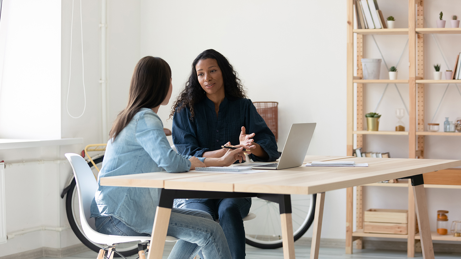 Smiling african american woman communicating with caucasian female colleague, sitting together at table in modern office. Young mixed race coworkers discussing working issues together at workplace.